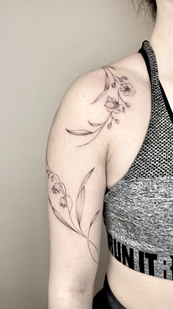Filigranes florales Schultertattoo in Fineline-Stil mit Blüten und Blättern, gestochen auf dem Oberarm einer Frau.

