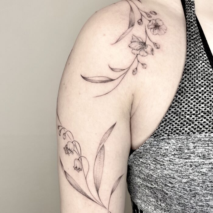 Filigranes florales Schultertattoo in Fineline-Stil mit Blüten und Blättern, gestochen auf dem Oberarm einer Frau.