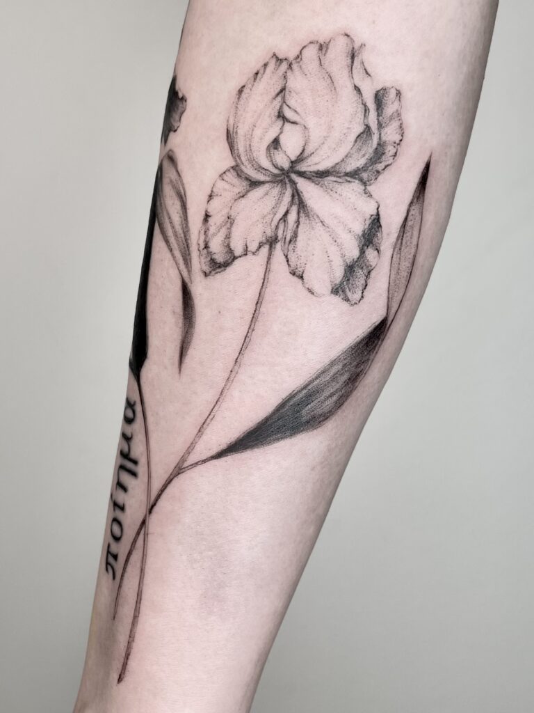 Feine Tattoo-Arbeit einer Irisblume auf dem Unterarm einer Frau, gestochen in einem realistischen Fineline-Stil. Die Blütenblätter wirken weich schattiert, während ein Schriftzug am Arm vertikal verläuft. Cover-up-Tattoo, aufgenommen in einem Studio in Zürich.