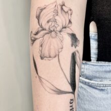 Detailliertes Fineline-Tattoo einer Irisblüte auf dem Oberarm einer Frau, mit sanften Schwarz-Grau-Schattierungen. Die Blume wurde über ein älteres Motiv gestochen (Cover-up) und zeigt eine elegante, feminine Linienführung. Tätowiert in einem Zürcher Studio. fineline tattoos in zürich