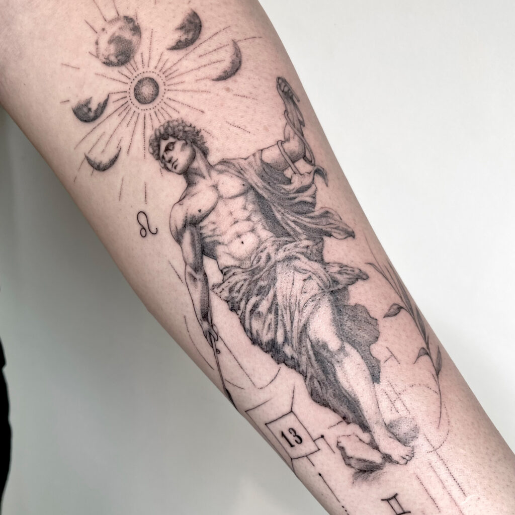 Fein gestochenes Schwarz-Weiß-Tattoo auf einem Unterarm: eine antik wirkende männliche Statue mit Tuch, umgeben von Sonne, Mondphasen und astrologischen Symbolen.