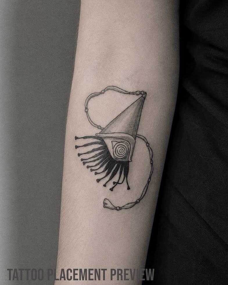 Fineline Tattoo mit Brimmed Hat aus Witch Hat Atelier / Atelier of Witch Hat, spitzem Zauberhut, Auge Symbol, schwarzen Fransen, Kordel und kleinen Perlen. Black and Grey Manga Tattoo auf dem Unterarm.